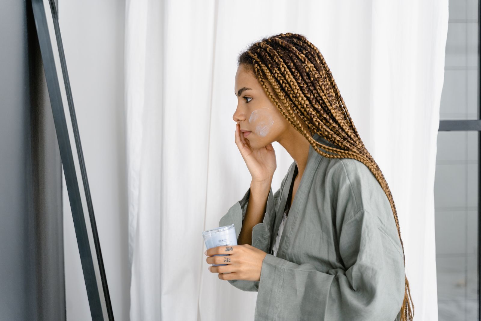 A woman applies white cream to her cheek while holding a jar beside a mirror
