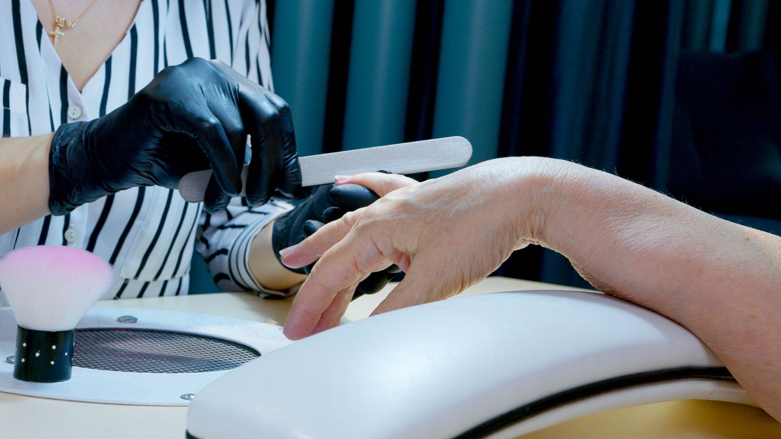 A gloved manicurist holds a manual nail file against the edge of a client's fingernail