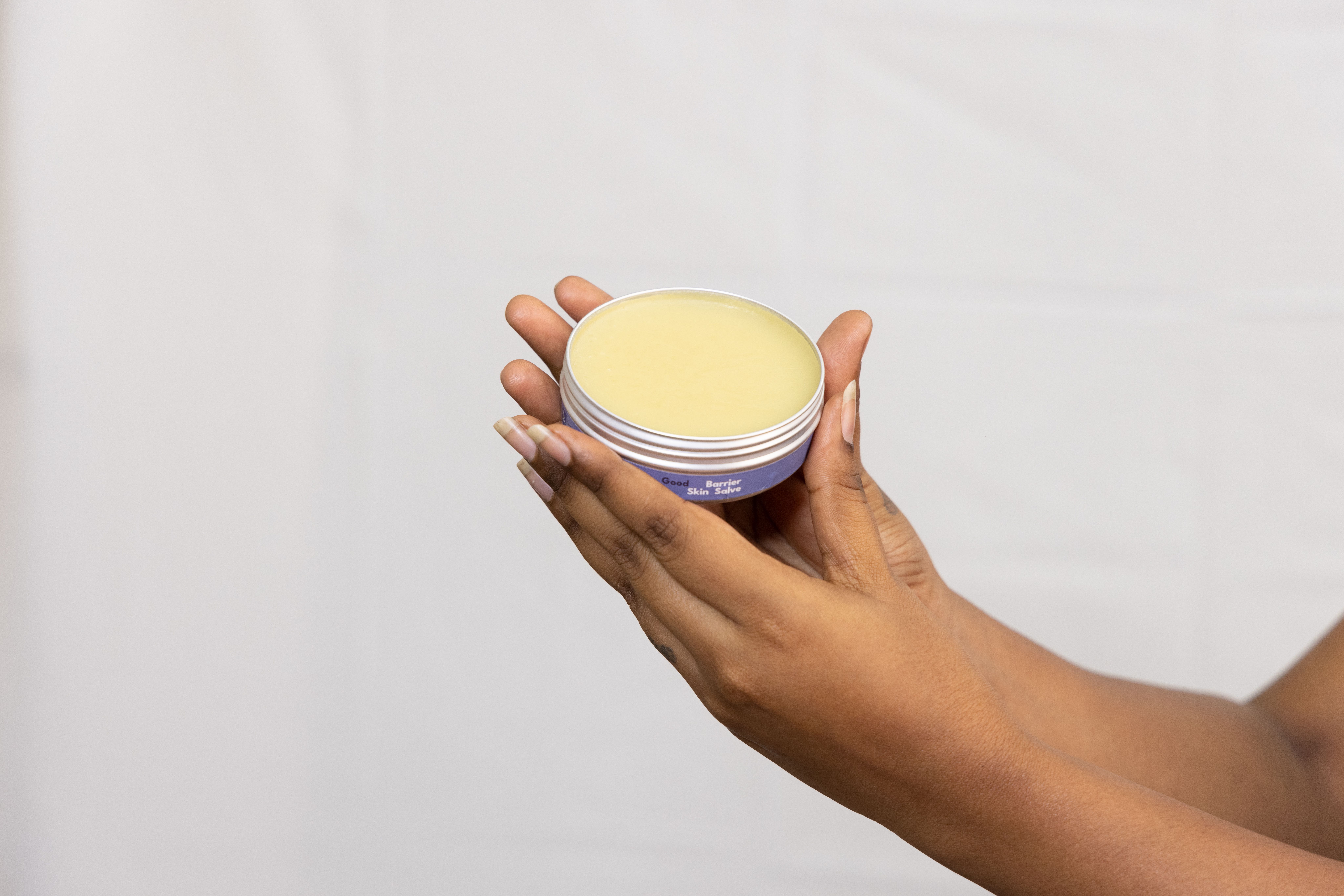Hands holding an open cosmetic salve tin against a pale background
