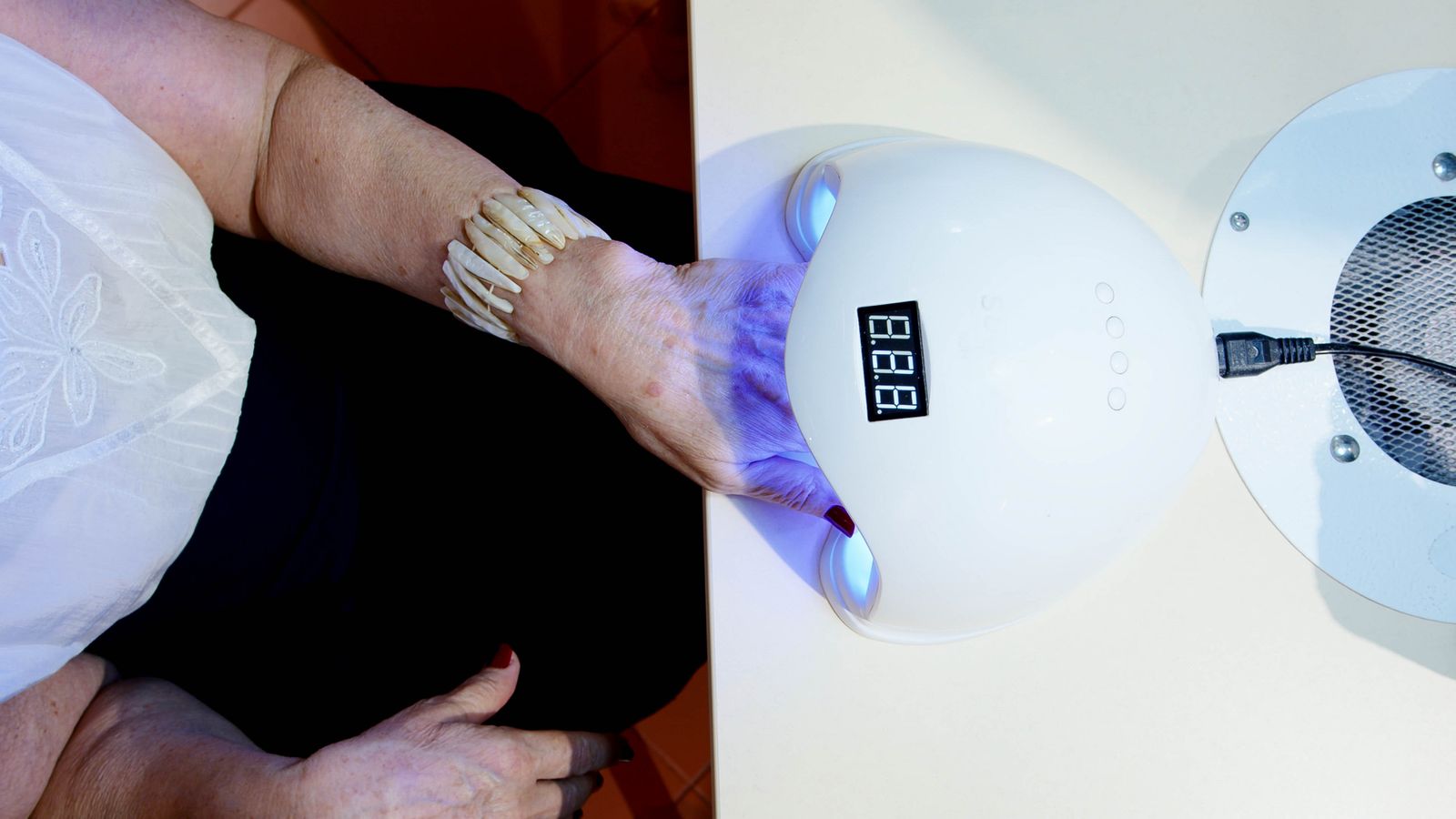 A hand with red nail polish rests inside a white nail lamp on a salon table