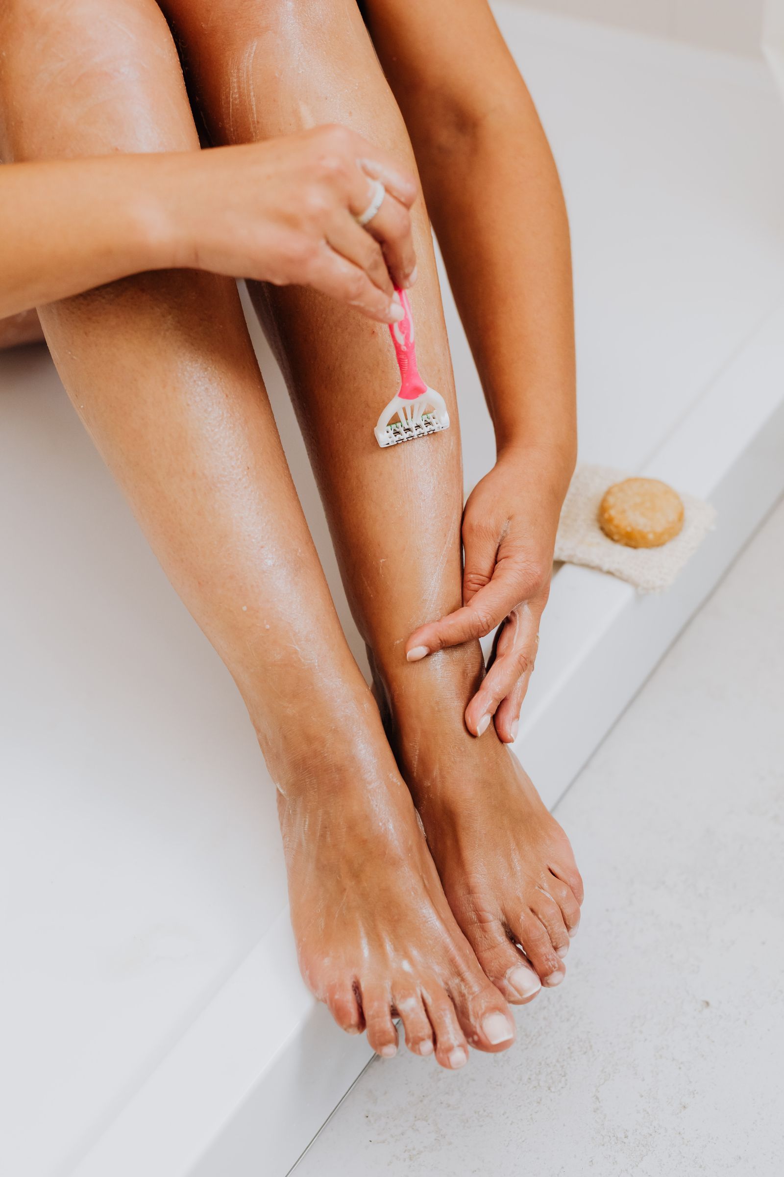 A seated person uses a pink manual razor on a lower leg with visible shaving lather beside a white bathtub