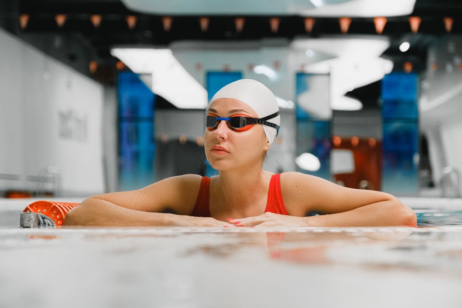 A swimmer in a white cap and goggles rests her arms on an indoor pool edge
