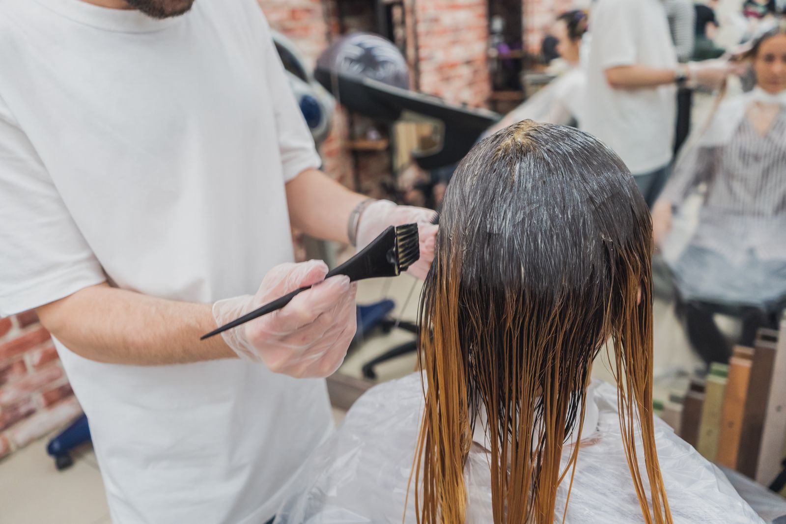 A gloved stylist holds a color brush beside a client's coated hair in a salon